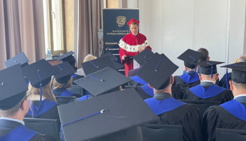 Rektor uniwersytetu, ubrany w czerwoną długą togę z kołnierzem, składa życzenia absolwentom.